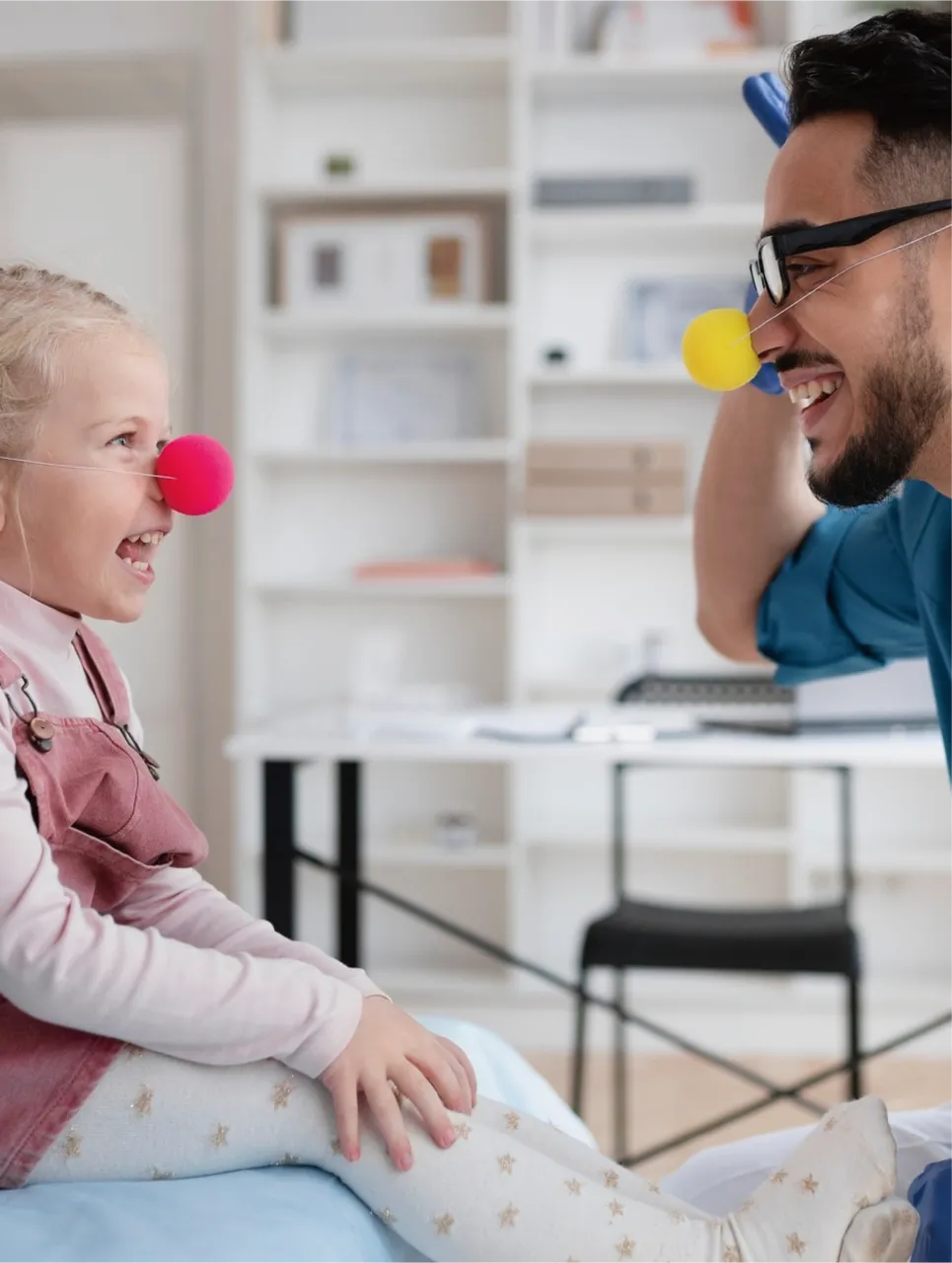 A pediatrician and a child wearing clown nose, smiling at one another